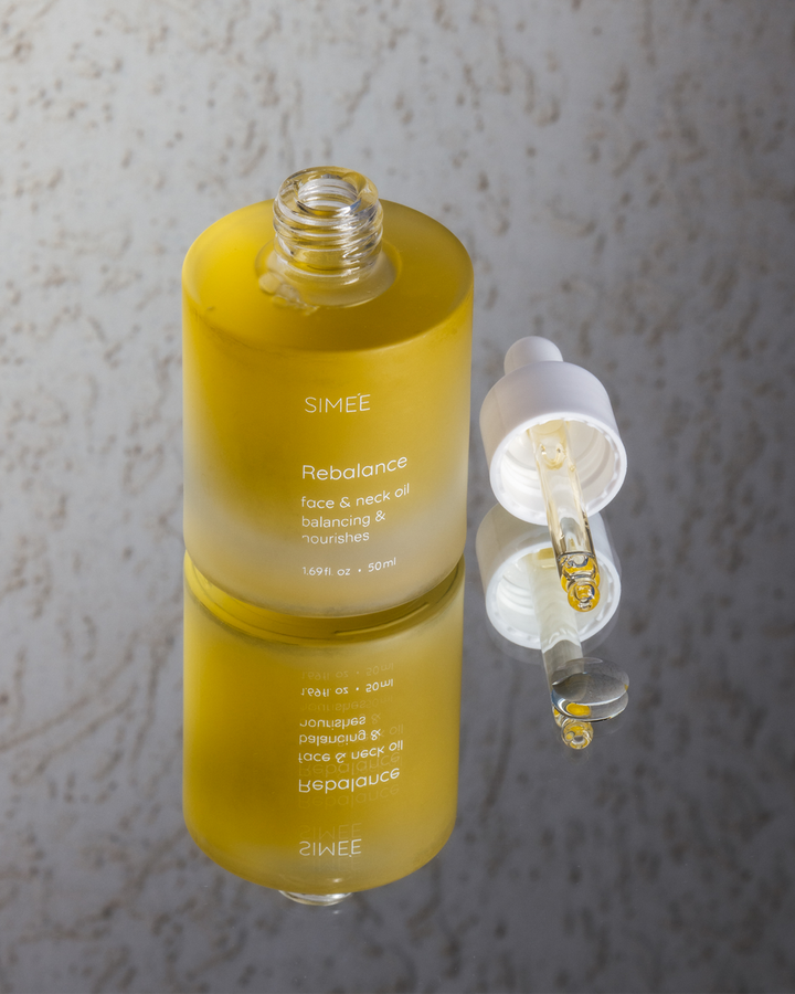 Open bottle of SIMÉE Rebalance face oil with a dropper and a drop of oil suspended over a marbled surface